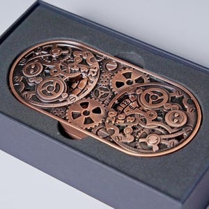 May include: A copper-colored, steampunk-style belt buckle featuring an intricate design of gears and cogs. The rectangular buckle is presented in a black box, highlighting the detailed mechanical artwork. The buckle's design includes various sizes of gears.