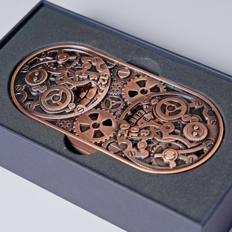 May include: A copper-colored, steampunk-style belt buckle featuring an intricate design of gears and cogs. The rectangular buckle is presented in a black box, highlighting the detailed mechanical artwork. The buckle's design includes various sizes of gears.