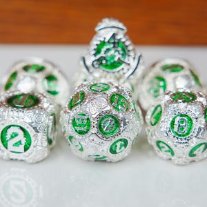 May include: A set of six polyhedral dice featuring a metallic silver finish with intricate gear-like textures and vibrant green enamel inlays. Each die displays clear, raised numbers in a bold font, showcasing a mechanical design aesthetic.