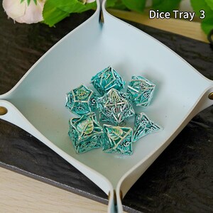 May include: A pale blue leather dice tray, square with rounded corners, holding several teal metal dice. The dice feature detailed leaf and geometric patterns. The text "Dice Tray 3" is visible. The tray is approximately 20 cm across.