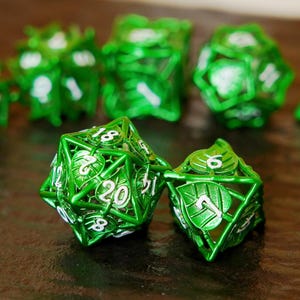 May include: A collection of green metal dice with white numerals. The dice feature a leaf design and are set against a dark backdrop. These dice are for tabletop games.