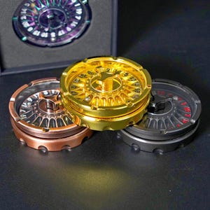 May include: A collection of metallic fidget spinners in gold, bronze, and black. Each spinner features a roulette wheel design with numbers and a central bearing. One spinner is displayed inside a black box.