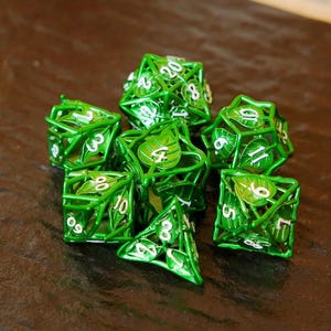 May include: A set of seven green metal dice with a leaf design. Each die has white numbers and a unique shape, including a D4, D6, D8, D10, D12, D20, and a second D10. The dice are arranged on a brown surface.