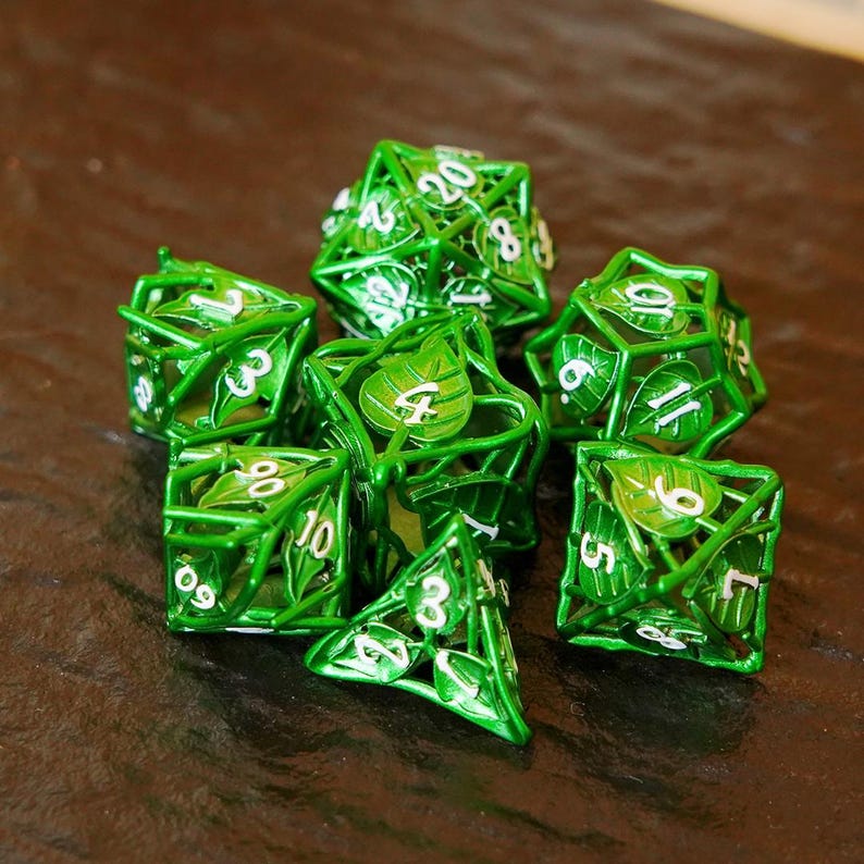 May include: A set of seven green metal dice with a leaf design. Each die has white numbers and a unique shape, including a D4, D6, D8, D10, D12, D20, and a second D10. The dice are arranged on a brown surface.