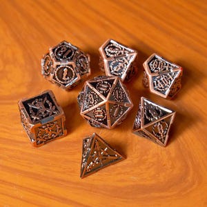 Könnte beinhalten: Ein Set aus sieben Metalldürfeln in verschiedenen geometrischen Formen, darunter ein W20, W12, W10, W8, W6, W4 und ein W20. Die Würfel haben eine Bronze- und Schwarz-Oberfläche mit aufwendigen Designs und Zahlen. Die Würfel liegen auf einer Holzoberfläche.