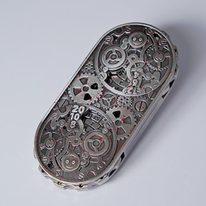 May include: A silver-colored, rectangular mechanical device with intricate gear and cog designs. The surface features the numbers "20 10 8" and red accents. The device has a complex, industrial aesthetic.