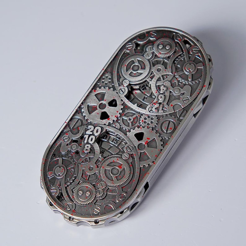 May include: A silver-colored, rectangular mechanical device with intricate gear and cog designs. The surface features the numbers "20 10 8" and red accents. The device has a complex, industrial aesthetic.