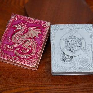 Könnte beinhalten: Zwei rechteckige Metallgehäuse mit Klappdeckeln. Das linke Gehäuse ist metallisch rosa mit einem Relief eines Drachen und Zahnradmustern. Das rechte Gehäuse ist silberfarben mit einem Relief eines kreisförmigen Uhrwerkmechanismus.