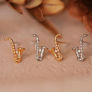 May include: Three gold and silver saxophone earrings with a single clear rhinestone on each. The earrings are on a light brown surface.
