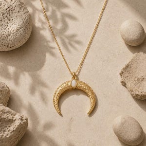 May include: A gold-toned necklace featuring a crescent moon pendant with an intricate filigree pattern and a small, oval-shaped white stone set at the center. The pendant hangs from a delicate gold-toned chain, creating a balanced and detailed piece.