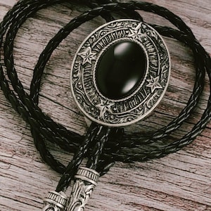 Può includere: Una cravatta a bolo nera con un ciondolo ovale color argento con un centro in pietra nera e dettagli a stella. Il ciondolo è collegato a un cordino in pelle nera intrecciata con punte decorative argentate. La cravatta è esposta su una superficie di legno.