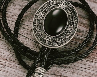 Könnte beinhalten: Eine schwarze Bolo-Krawatte mit einem silberfarbenen ovalen Anhänger mit einem schwarzen Stein in der Mitte und Sternakzenten. Der Anhänger ist mit einer geflochtenen schwarzen Lederschnur mit dekorativen silbernen Spitzen verbunden. Die Krawatte wird auf einer Holzoberfläche präsentiert.