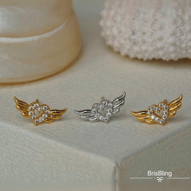 Puede incluir: Dos pares de pendientes con un dise&ntilde;o de coraz&oacute;n y alas. Un par es dorado con cristales transparentes y el otro par es plateado con cristales transparentes.