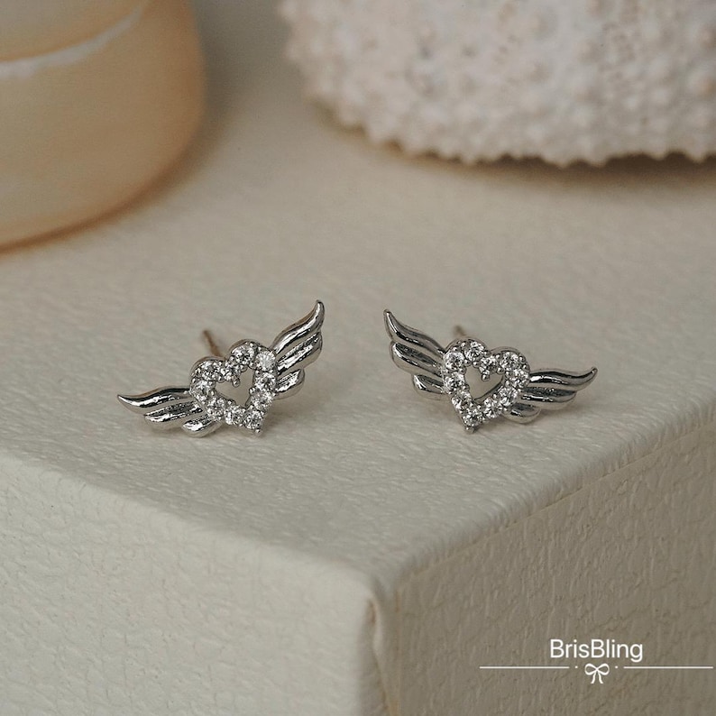 Puede incluir: Un par de pendientes de plata con un dise&ntilde;o de coraz&oacute;n y alas. El coraz&oacute;n est&aacute; lleno de peque&ntilde;os cristales transparentes.
