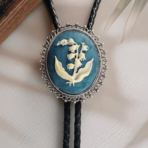 Pode incluir: Gravata bolo oval com fundo azul e design de lírio-do-vale branco. O pendente é emoldurado por uma filigrana prateada. O cordão de couro preto é trançado.