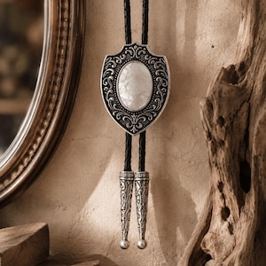 Op de afbeelding: Een bolo-das met een schildvormige hanger van zilverkleurig metaal met ingewikkelde krulmotieven en een centrale ovale witte steen. De hanger is bevestigd aan een zwart gevlochten koord dat eindigt in twee bijpassende zilverkleurige metalen punten.
