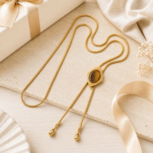 May include: A gold-toned bolo tie necklace featuring a long, flexible snake chain. The centrepiece is a diamond-shaped gold metal plaque with an oval, brown tiger's eye gemstone. The two ends of the chain are finished with smooth, gold-toned teardrop charms.