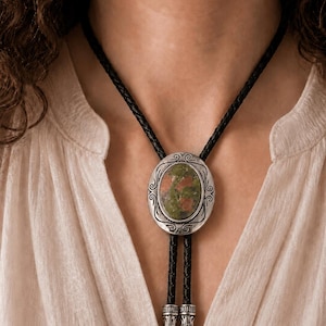 May include: A bolo tie featuring a central oval-shaped stone with mottled green and reddish-brown hues, set within an ornate silver-toned metal frame. The tie includes a black braided leather cord finished with two matching silver-toned metal tips.