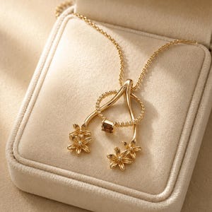 Puede incluir: Un collar de oro con un colgante en forma de horquilla adornado con dos pequeños racimos de flores en los extremos. Un anillo de oro fino con una pequeña piedra preciosa rectangular marrón cuelga del centro, unido a una delicada cadena de oro.