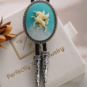 Può includere: Una cravatta a bolo con un unicorno alato color crema su uno sfondo ovale turchese, incorniciato da un motivo a corda argentata. Ha un cordino in pelle nera e punte argentate decorative. La cravatta è su una scatola bianca.
