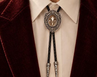 Könnte beinhalten: Eine Bolo-Krawatte mit einem silberfarbenen, schildförmigen Mittelstück und einem geprägten Fleur-de-Lis-Emblem. Die Krawatte besteht aus einer geflochtenen schwarzen Lederschnur, die mit zwei passenden silberfarbenen Metallspitzen mit Gravur endet.