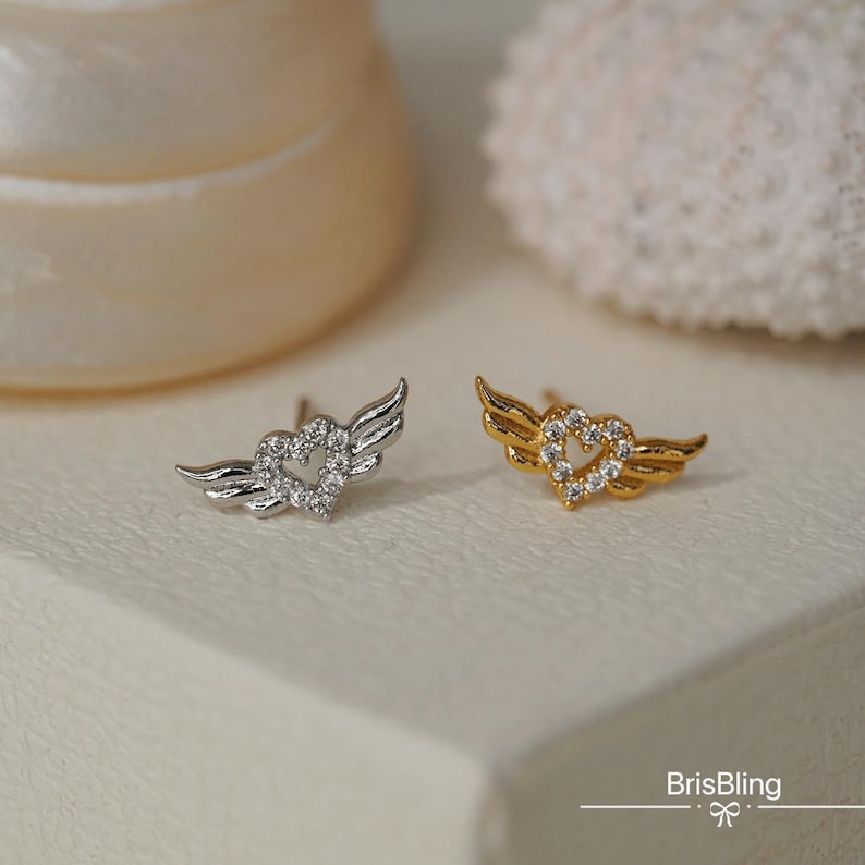 Puede incluir: Un par de pendientes con un dise&ntilde;o de coraz&oacute;n con alas. Un pendiente es de plata con cristales transparentes, y el otro es de oro con cristales transparentes.