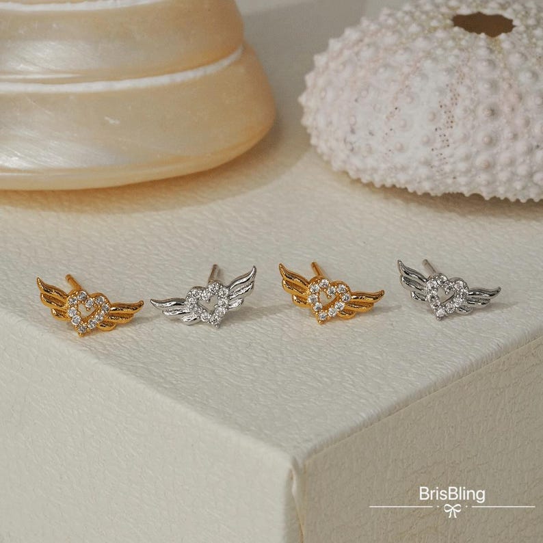 Puede incluir: Tres pares de pendientes con un dise&ntilde;o de coraz&oacute;n y alas. Los pendientes est&aacute;n hechos de metal plateado y dorado y tienen piedras de circonita c&uacute;bica transparentes.