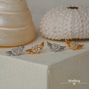 Puede incluir: Tres pares de pendientes con un dise&ntilde;o de coraz&oacute;n y alas. Los pendientes est&aacute;n hechos de metal plateado y dorado y tienen cristales transparentes.