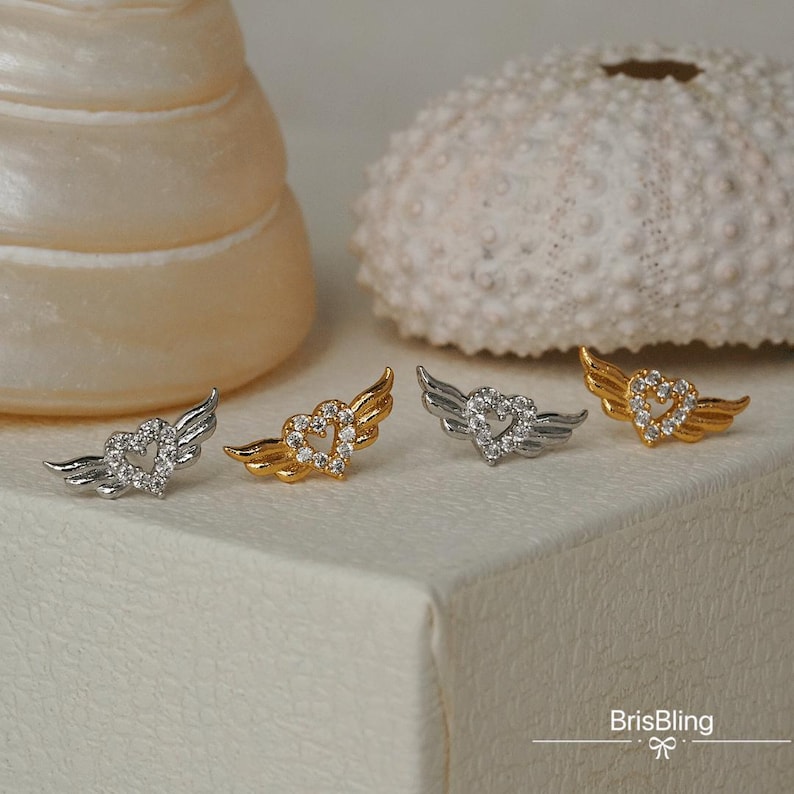 Puede incluir: Tres pares de pendientes con un dise&ntilde;o de coraz&oacute;n y alas. Los pendientes est&aacute;n hechos de metal plateado y dorado y tienen cristales transparentes.