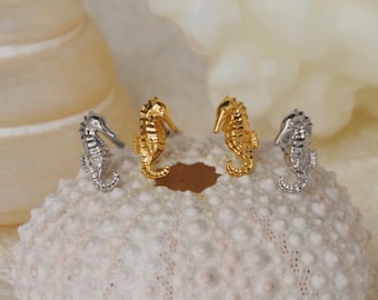 May include: A pair of gold and silver seahorse stud earrings. The earrings are displayed on a white seashell background.
