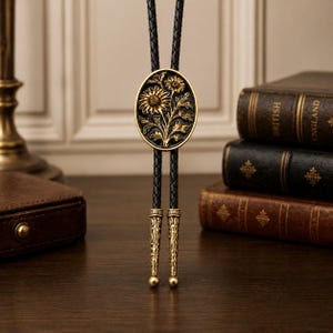 May include: A bolo tie featuring a black braided leather cord and an oval-shaped gold-toned metal centerpiece with a raised floral design. The cord ends are finished with matching gold-toned metal tips that have an intricate engraved pattern.
