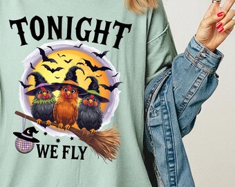 May include: A light green t-shirt with the text "TONIGHT WE FLY" above a graphic of three birds wearing witch hats, perched on a broomstick, with a full moon and bats in the background. The shirt also has the text "Bay Comfort Colors 1717".