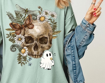 May include: A light green t-shirt featuring a skull design with flowers, a butterfly, and a ghost. The text "Bay Comfort Colors 1717" is printed on the upper right. The shirt has short sleeves and a relaxed fit.