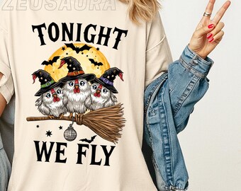 May include: Ivory-colored t-shirt with the text "TONIGHT WE FLY". The shirt features a graphic of three owls wearing witch hats, perched on a broomstick, with a full moon and bats in the background. The shirt is labeled "Comfort Colors 1717".