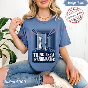 May include: A blue Gildan 5000 t-shirt featuring a chess-themed graphic. The graphic includes a queen and pawn, with the text "THINK LIKE A GRANDMASTER". The shirt also has the text "Indigo Blue" and a "Do Not Copy" stamp.