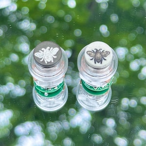 Op de afbeelding: Twee doorzichtige, cilindrische flessenstoppers met bijenontwerpen. Eén stopper heeft een grijze bij, de andere een bruine bij. Elke stopper heeft een groene band in het midden.