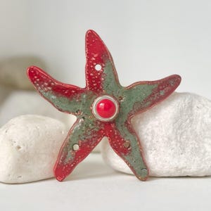 May include: A red and green starfish-shaped decorative object with a red center. The starfish has a textured surface and is resting on white stones. The object has a hole in each of its five points.