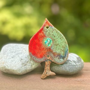 May include: A heart-shaped ceramic ornament featuring a vibrant red and green glazed finish with speckled details. The center contains a circular iridescent abalone shell inlay. A small hole is positioned at the top for hanging, and a stem extends from the bottom.