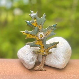 Può includere: Spilla in ceramica a forma di foglia, a tema natura. La spilla presenta un design dettagliato con tonalità giallo e verde-blu, e un piccolo emblema rotondo con l'immagine di una foglia. La spilla è appoggiata su una pietra bianca su uno sfondo verde sfocato.