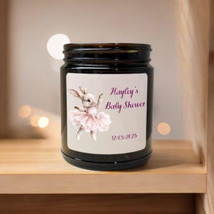 May include: A dark brown glass jar with a white label featuring a cartoon bunny in a pink tutu. The label reads "Hayley's Baby Shower" and the date "12/05/2025". The jar is on a wooden shelf.