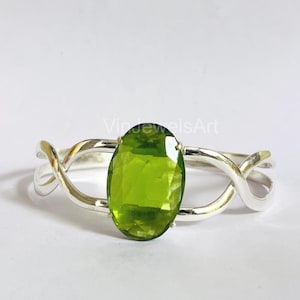 Puede incluir: Un anillo de plata con una gema grande, ovalada y verde claro. La banda tiene un diseño único e entrelazado. El anillo está sobre un fondo blanco. El texto "VindjewelsArt" es visible en el fondo.