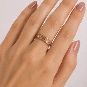 May include: A rose gold band ring featuring a continuous embossed floral pattern with small flowers and swirling vine details. The ring has a flat profile and a polished finish, encircling the finger with a repeating botanical design.