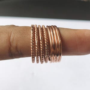May include: A selection of copper-coloured rings, some with a twisted rope design, stacked on a finger. The rings vary in style, with some smooth and others textured. The rings are a warm, metallic colour.