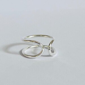 May include: A silver ring with a unique, open design. The ring features a continuous, flowing shape with two circular elements and a curved band. The polished metal reflects light, creating a bright and modern aesthetic. The ring is set against a plain white background.