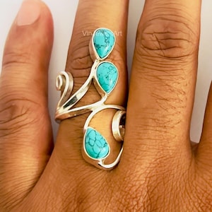 Puede incluir: Un anillo de plata con tres piedras de turquesa en forma de lágrima con vetas oscuras. La banda de metal presenta un diseño abierto y en espiral que se envuelve alrededor del dedo, mostrando un acabado pulido y un color azul verdoso.