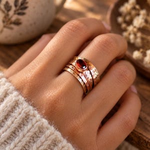 Könnte beinhalten: Ein breiter Mehrfachring aus kupferfarbenem Metall mit gehämmertem Muster. Das mittlere Band ist mit einem ovalen, in einer Zargenfassung sitzenden Granat-Edelstein in tiefem Rot besetzt. Das Design besteht aus mehreren miteinander verbundenen Ringen.