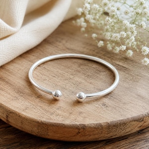 Op de afbeelding: Een gepolijste zilveren armband met een glad, open ontwerp en een kleine, bolvormige kraal aan elk uiteinde. De metalen band heeft een reflecterende afwerking en een cirkelvormige structuur, wat zorgt voor een strak en eenvoudig uiterlijk.