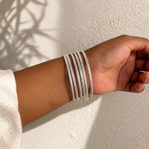 Puede incluir: Un conjunto de cinco brazaletes finos de tono plateado usados en la muñeca. Cada brazalete presenta una superficie texturizada y martillada con un patrón repetitivo de pequeñas hendiduras circulares que captan la luz, creando un aspecto metálico.