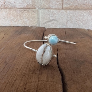 Puede incluir: Una pulsera de plata con una concha blanca y una gema azul claro. La pulsera está colocada sobre una superficie de madera, con una pared de ladrillo al fondo. La joya está diseñada con un estilo minimalista.