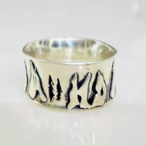 May include: A silver ring with a mountain and tree design. The ring has a wide band with a textured surface. The mountain and tree design is cut out of the band, creating a unique and detailed look. The ring is likely made of sterling silver.